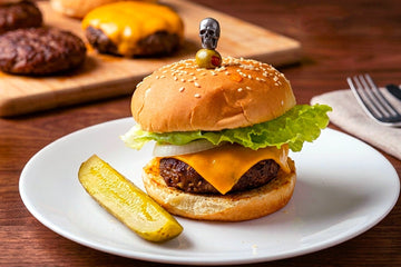 a cheeseburger with lettuce and a sesame seed bun on a white plate with a pickle.