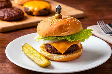 a cheeseburger with lettuce and a sesame seed bun on a white plate with a pickle.