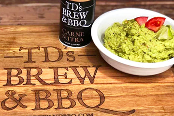 a white bowl with guacamole tomatoes and lime sitting on a wood cutting board with TD's Brew & BBQ burned into it and a bottle of Carne Sutra seasoning.