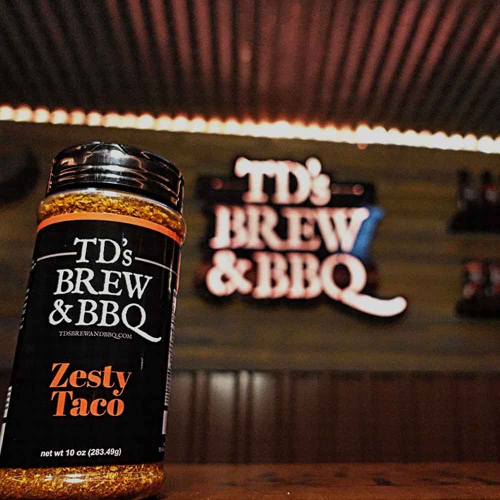 Jar of TD's Brew & BBQ Zesty Taco seasoning in front of a neon sign.