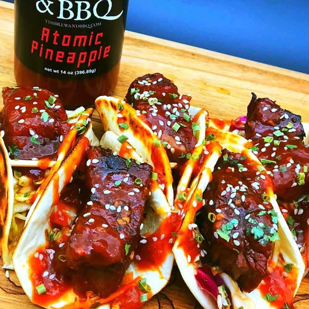 Saucy pork belly on pita with sauce and garnishes on a wooden board with a bottle of Atomic Pineapple sauce