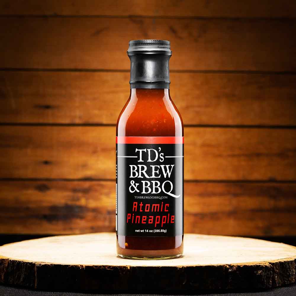 Bottle of TD's Brew & BBQ Atomic Pineapple sauce on a wooden surface with a wooden background