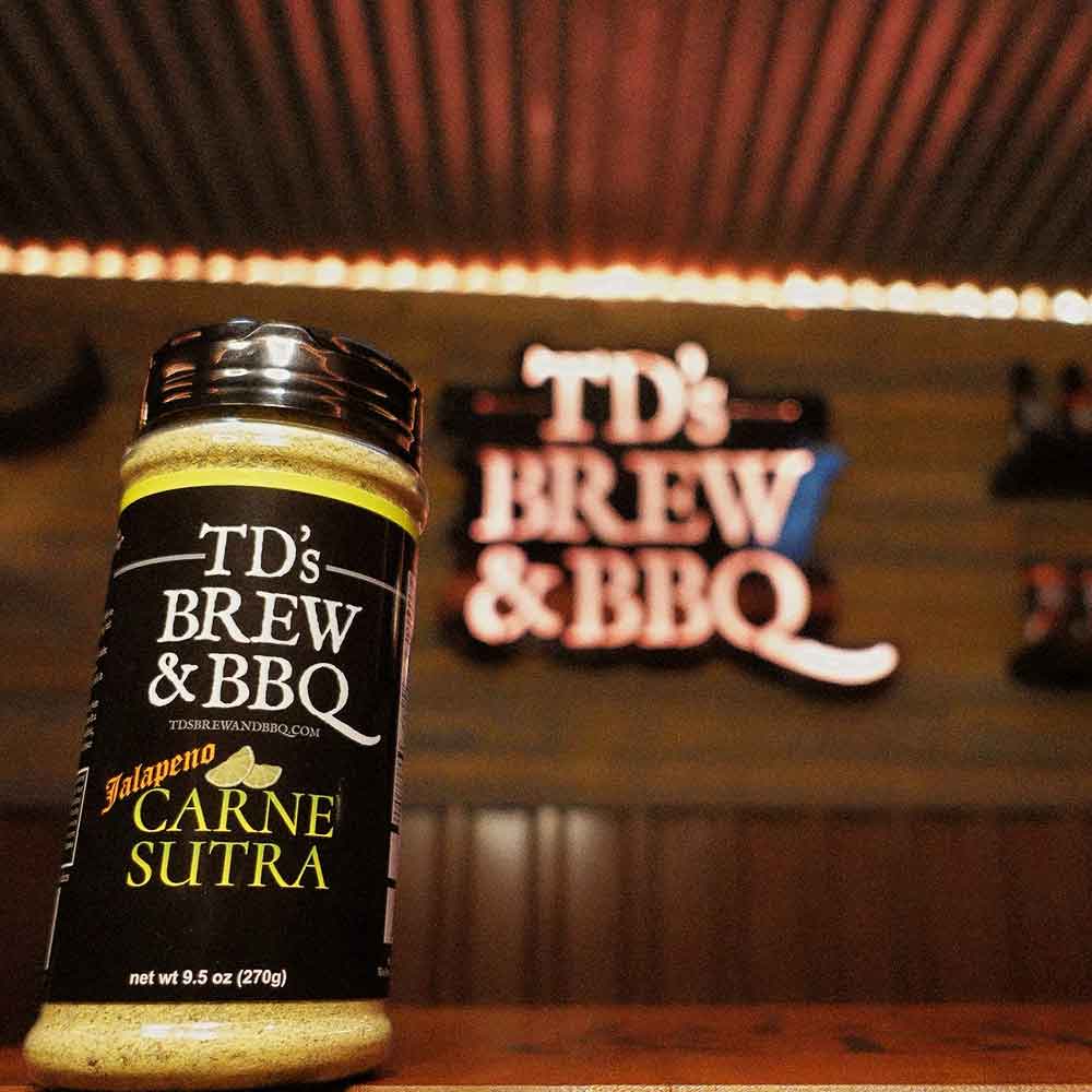 Jar of TD's Brew & BBQ Carne Sutra seasoning with a blurred background featuring 'TD's Brew & BBQ' sign.