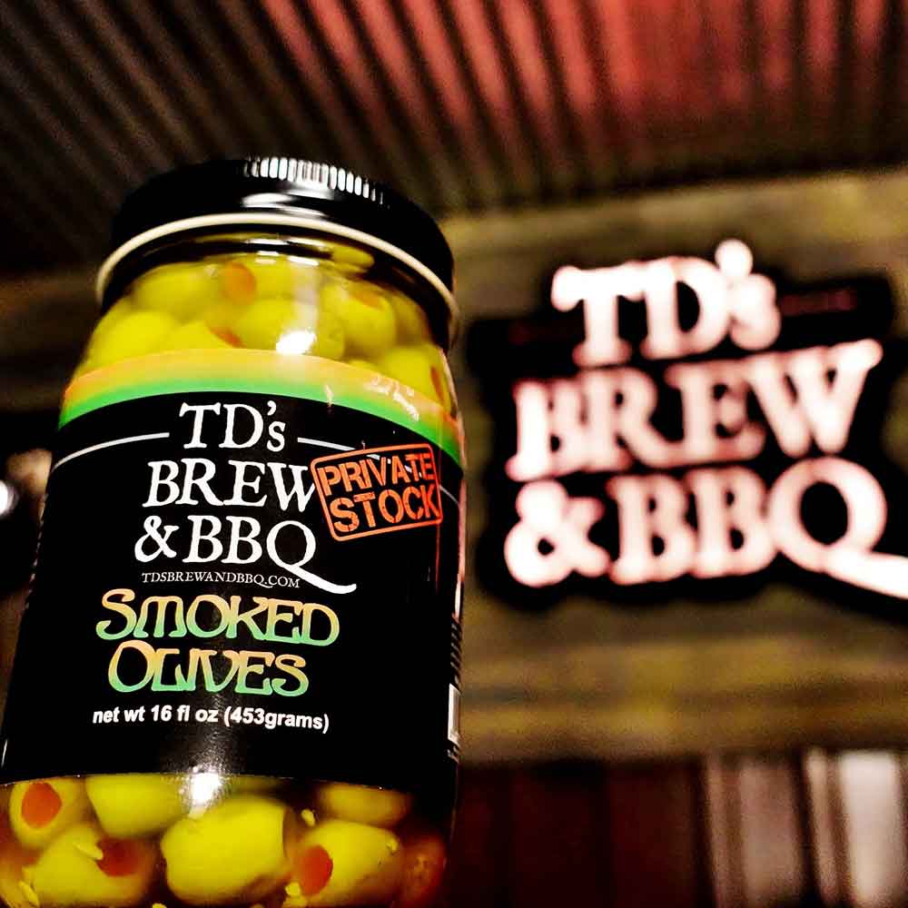 Jar of TD's Brew & BBQ Smoked Olives on a counter with a neon sign behind
