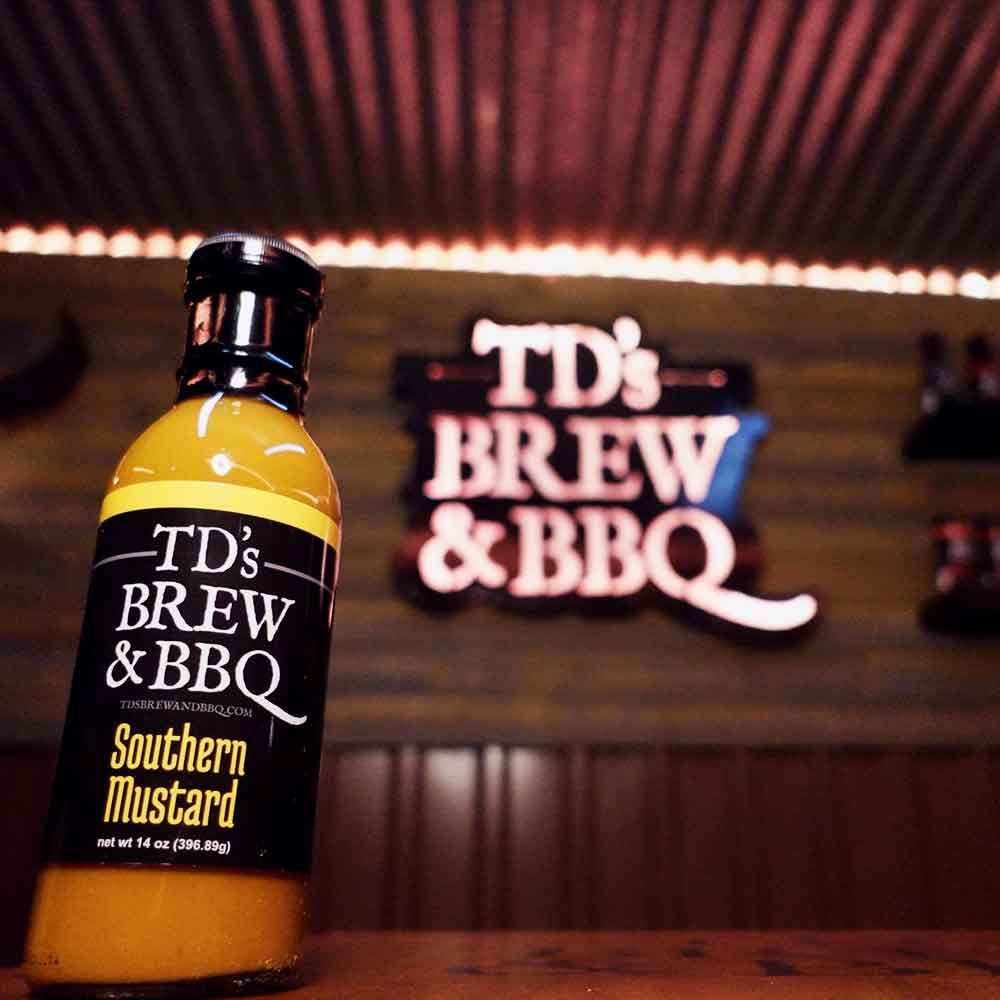 Bottle of TDs Brew and BBQ Southern Mustard Sauce on a wood shelf with neon sign behind