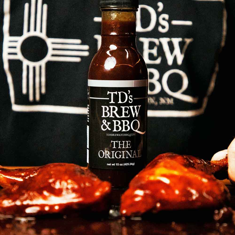 Bottle of TD's Brew & BBQ sauce with ribs in the foreground and a branded shirt in the background.